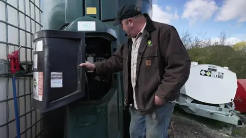 Dan Willis opens a tank to check how much red diesel he has left. There is a machine behind him. He is wearing a brown jacket and a cap. He is facing away from the camera.