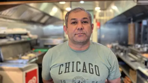 A man with grey hair and a green T-shirt branded Chicago stands in the kitchen area of a takeaway food outlet facing the camera with his hands behind his back