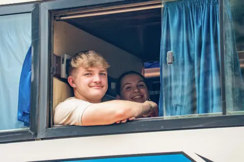 Studio Lambert/BBC Fin and Sioned look out of the window of a bus at the camera. Fin has his arm resting on the bottom of the open window, with blue curtains to the right. He has short blond hair. Sioned can be seen sitting next to him.