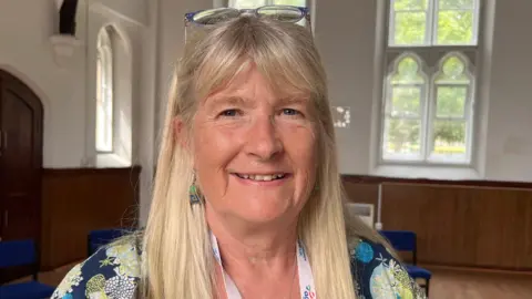 A woman with long blonde hair smiling, with glasses on her head, wearing a white lanyard and a navy floral t-shirt, with a white wall and windows in the background.