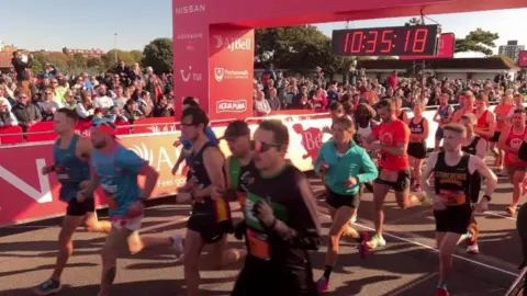 BBC Crowds watching on as runners cross the start line with red barriers along the road and a digital clock in the background