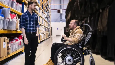 Sky Documentaries Daniel Radcliffe and David Holmes. Daniel is standing. David is in a wheelchair looking up at Daniel. The background is of a warehouse.