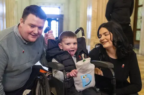 PA Media Dáithí Mac Gabhann sits in his wheelchair, holding a hot water bottle as his parents Máirtín and Seph look on