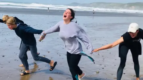 A young women in a grey jumper, laughing as she holds hands with people on the beach