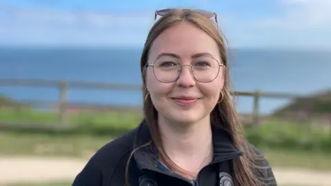 A woman wearing glasses and a blue fleece is standing in front of a fence. The sea is visible in the background. 