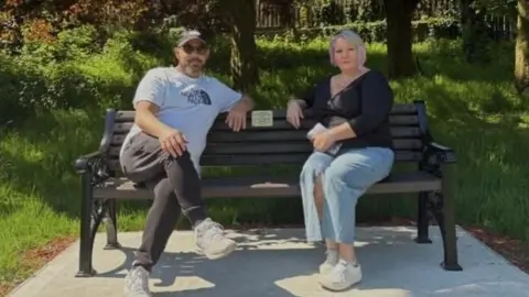 Leanne Booth Leanne on bench in next to her husband on sunny day. She wears a black cardigan and trousers and he wears a white t-shirt and black jeans and sunglasses.