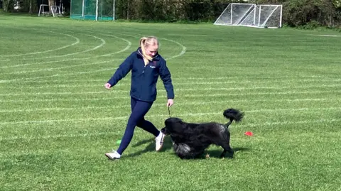 Sam Read/BBC Megan is pictured running with a dog on a lead on a sports pitch. She has long blonde hair and is wearing a blue tracksuit. The dog is black and fluffy and has a patch of white hair on its chest.