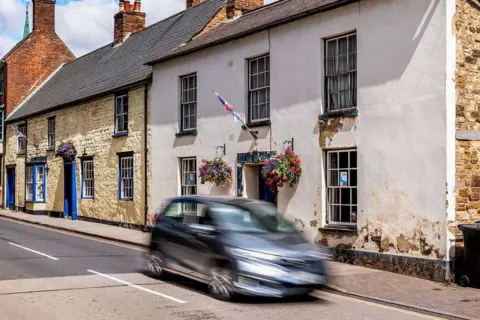 National Highways A long exposure image of a blurred grey car travelling past properties in the centre of Towcester.