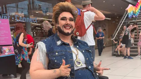 Harriet Morter/BBC Drag King Crip Ladywood. They are sitting in a wheelchair in The Forum in Norwich and are looking directly at the camera and smiling. They have short voluminous hair and are wearing a denim vest top with sparkly tassels coming off the sleeves. The outfit has rhinestones on it. 