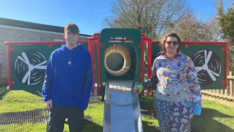 Daniel Jackson (left) and Jess Ashbridge (right) stand either side of the Lancaster bomber climbing frame. The green climbing frame has 'wings' bordered in red with propellers painted on. A slide comes out from the front of the page, after a wooden crawl space. Daniel is smiling and wearing a dark blue hoody. Jess is smiling and wearing rainbow-coloured sunglasses, a hoodie with rainbows and emojis and brightly patterned trousers. 