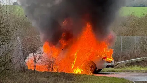 A silver BMW is parked on a road and engulfed in bright orange flames.