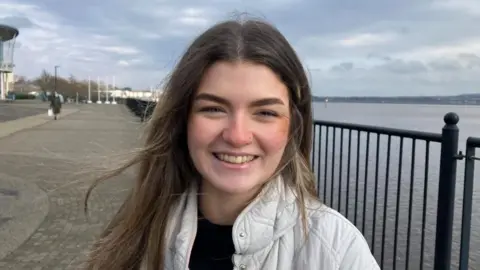 A woman with long dark hair is standing on a quayside in front of a river. She is wearing a white jacket over a black top.
