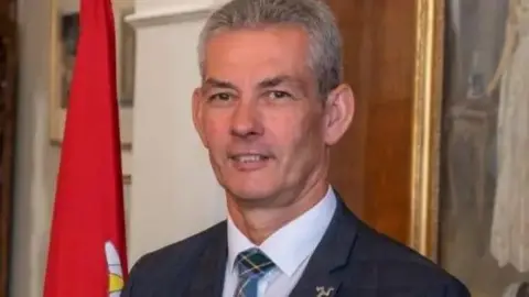 Tim Crookall, a man with grey hair wears a suit, there is a red flag in the background.