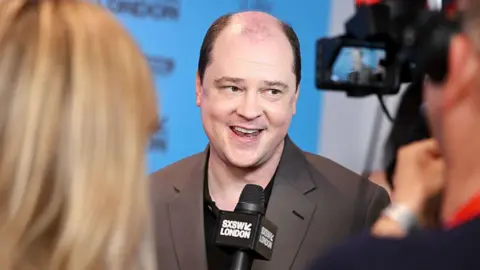 Getty Images Mike Flanagan pictured mid-interview on a red-carpet. He's wearing a dark brown suit jacket over a black t-shirt. The camera focuses on him over the shoulders of a reporter and cameraman.