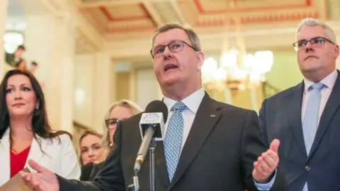 Getty Images A man speaks into a microphone. He has grey hair and is wearing glasses. His arms and hands are stretched out. He is wearing a dark suit, white shirt and light blue patterned tie. He has a fish badge pinned to his jacket. The man on his left is taller and has white hair. He is wearing glasses, a grey suit, light blue tie and white shirt. The woman on the left of the image has long dark hair. She is wearing a white jacket and red top underneath. The background is blurred.