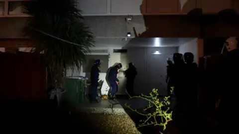 Three police officers outside a ground floor apartment at night time, dressed in helmets and riot gear, preparing to knock down the door