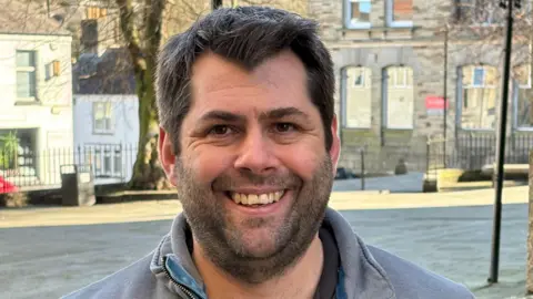 A man with a dark hair and a beard wearing a grey top smiling. 