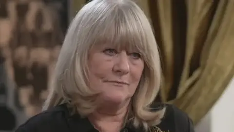 Christine Flack looks to the right of the camera during an interview. Her expression is serious and she is wearing a dark colored blouse. She has long blonde hair with fringes. 