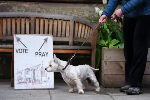 Jane Barlow / PA Media A white dog bows its head while on a lead by a sign with two arrows saying 'vote' and 'pray' in Inverleith, Edinburgh.