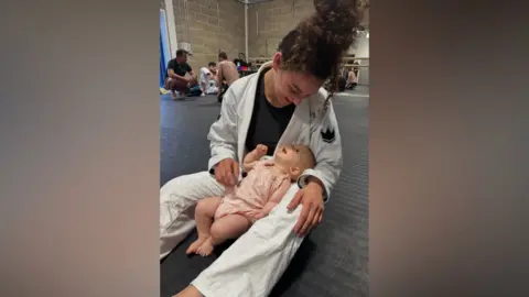 Bea Fernandes Bea Fernandes sits on a mat on the floor at a jiu-jitsu competition. She wears a white jiu-jitsu competition outfit. Her baby sits in the middle of her legs. Bea looks down at her baby and smiles while the baby is looking up and also smiling. 
