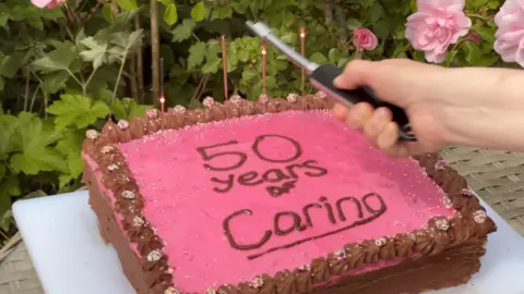 A hand holding a knife over a pink and brown cake with the inscription "50 years of caring". It is outside. Some pink roses can be seen blooming nearby.