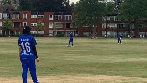 Louise Hulland/BBC The cricket ground with some players in their blue kit. Close to the camera is a woman with the name N.Amiri 16 on her shirt.