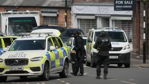 BBC Two armed police officers in bullet-proof vests and wearing black helmets stand next to three police cars and a police van on the street