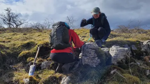 Graham Standring Two people with coats, hats and rucksacks plant in the countryside. There are some ragged rocks as well as grass and trees which have no leaves on them.