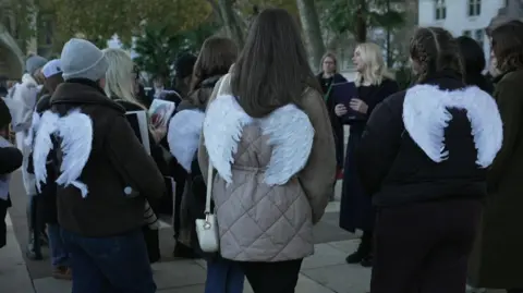 A number of women seen from behind wearing coats with small white angel wings on the back.