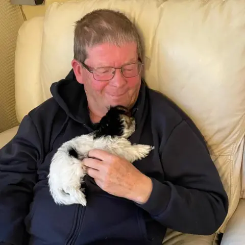 Siobhan Mackenzie Graham Mackenzie is an older man. He is wearing a blue fleece and sitting in an armchair with Gigi lying on his chest.