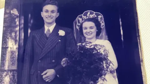 Sandra Beech A wedding photo of Iris and David Jefferies. They are standing arm in arm in front of a church.