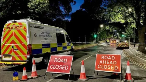 Police at the scene of a crash on Victoria Park Road, Leicester, at night with a cordon in place and road closure signs and cones