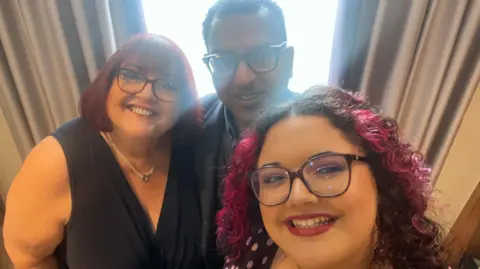 Ahmad A husband and wife stand behind their daughter, taking a selfie. The husband has dark skin, and his wife is Caucasian.