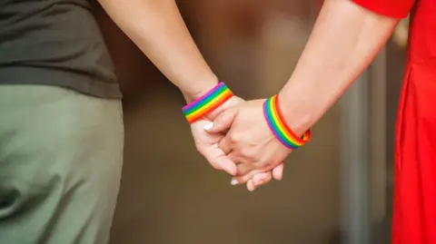 Getty Images Close up of two people holding hands wearing rainbow wrist bands