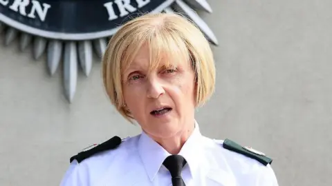 Barbara Gray wearing her PSNI uniform in front of the PSNI logo on the wall behind her. She has short blonde hair.