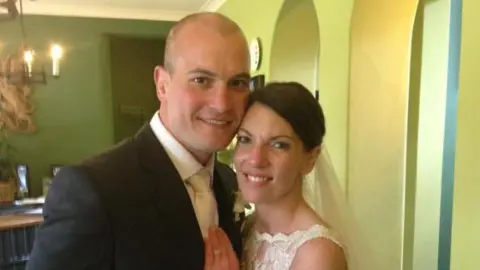 Image of a couple standing close on their wedding day. The man is wearing a black suit, white shirt and cream coloured tie. The bride has black hair tied in a bun and is wearing a veil and lace wedding dress. In the background can be seen a room with green walls and candle wall sconces.