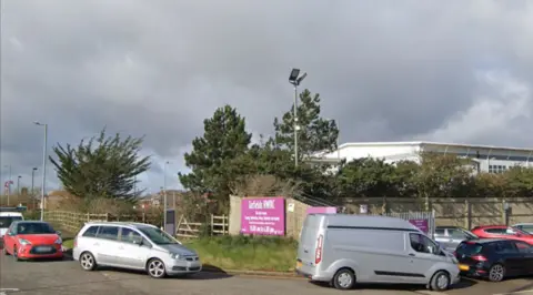 The Sixfields recycling centre, as detailed on a purple sign by the entrance. There is a queue of vehciles turning left into the site, including a red car, a grey MPV, grey van and a black Corsa. The site is fenced off with wooden fencing, with trees behind.