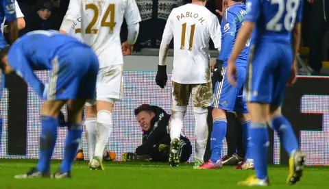 ANDREW YATES/AFP via Getty A boy in a black tracksuit lies on the ground by a football pitch's advertising hoarding with a pained expression on his face after a altercation with football players in blue and white kits standing around him.