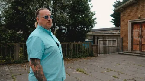 Neil Maggs Paul Holbrook wearing a blue shirt with his hands in his jeans on front of a building with a wooden fence outside