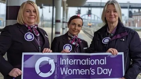 East Midlands Railway Sara Hardstaff, Sally Ashforth and Maria Alton holding an international womens day banner at Derby Railway Station