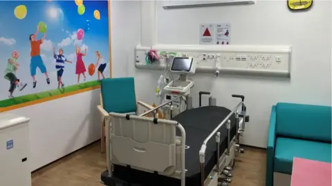 Worcestershire Royal Hospital Hospital room with a bed and a blue chair on the left hand side of it while a blue chair is to the right hand side. There is a picture of children playing with balloons on the wall