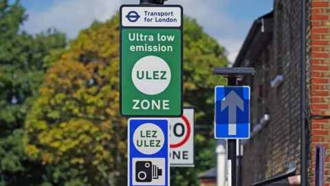 PA Media A green ULEZ sign and a blue LEZ and ULEZ sign with a camera on