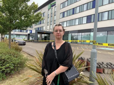 A woman with dark hair and glasses stands in front of yellow police tape in front of a large hotel building