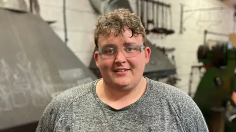 Ethan Lewis, a man wearing a grey top and glasses. He is stood in a blacksmith's forge.