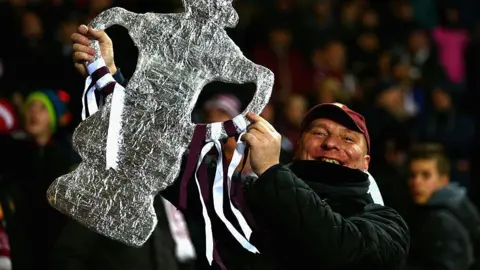Getty Images A Northampton Town fan holding up a foil replica of the FA Cup trophy. It has claret and white ribbons attached to it. The fan is wearing a claret baseball cap and has a broad smile