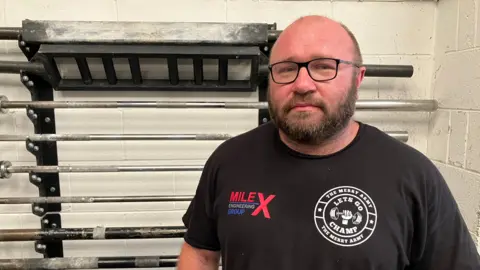 A man in a black T-shirt and glasses stands in front of a rack with multiple metal bars on it.