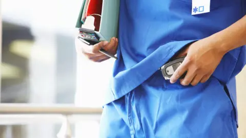 A doctor dressed in blue scrubs puts a pager on his trousers.
