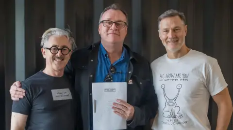 Channel 4 / Ben Blackall Davies stands in the middle of Cumming and Morrissey, with his hand around Cummings' shoulders. Davies holds the series script in one hand. Morrissey wears a white t-shirt with a graphic on, and has short grey hair. Cumming wears black round glasses and wears a black t-shirt. 