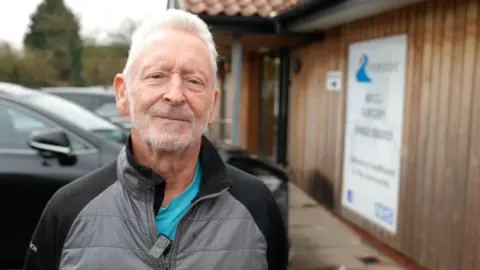 A man stands outside a surgery building on a wet walkway, wearing a grey and black jacket over a turquoise top. Behind him is the surgery’s wooden exterior, with a large sign displaying the Riverside logo next to the entrance. He has short grey hair and a closely cropped beard. Parked cars line the left side of the image and the building’s tiled roof is visible.