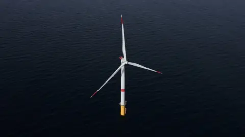 Reuters A wind turbine stands alone in a dark, calm ocean. The lower part of the tower, where it meets the water, is painted bright yellow, while the rest of the tower is white. At the top of the tower is a hub with three enormous blades attached, forming a shape like a giant three-pointed star. Each blade is long and slender, tapering toward the end. The ends of the blades have red markings.